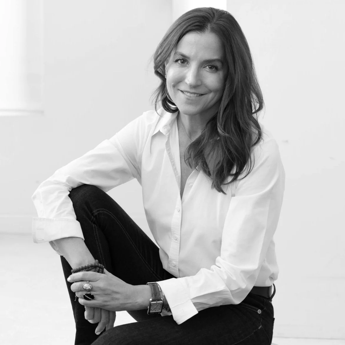 Black and white photo of Ann Watson, wearing a white shirt and black jeans, sitting on the floor with one knee up and her arm resting on it.