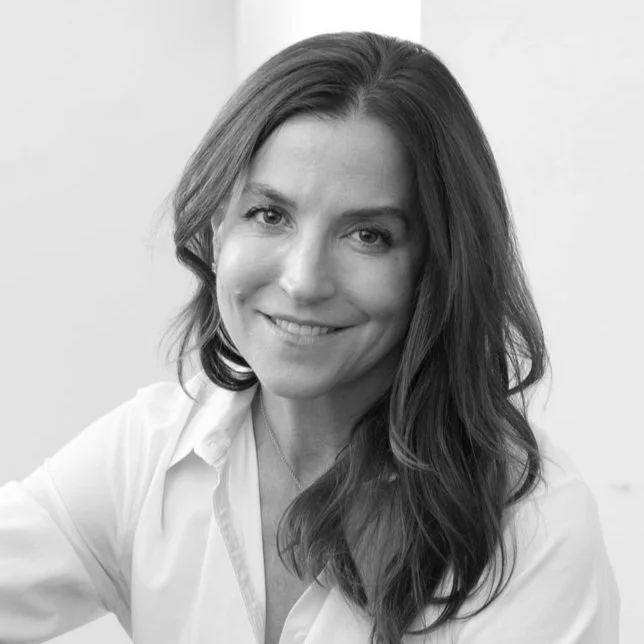 Black and white photo of Ann Watson, wearing a white shirt and black jeans, sitting on the floor with one knee up and her arm resting on it.