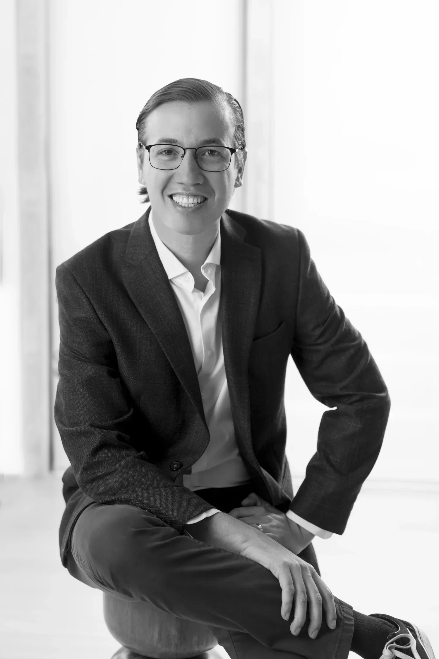 Black and white portrait of Adolfo Flores-Saiffe with glasses in a business suit, sitting on a ball, smiling at the camera.