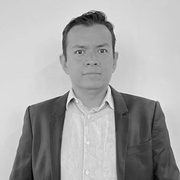 A black and white photograph of Carlos Campos in business attire, wearing a dark blazer and a white shirt, standing indoors by a white surface with a bright background.
