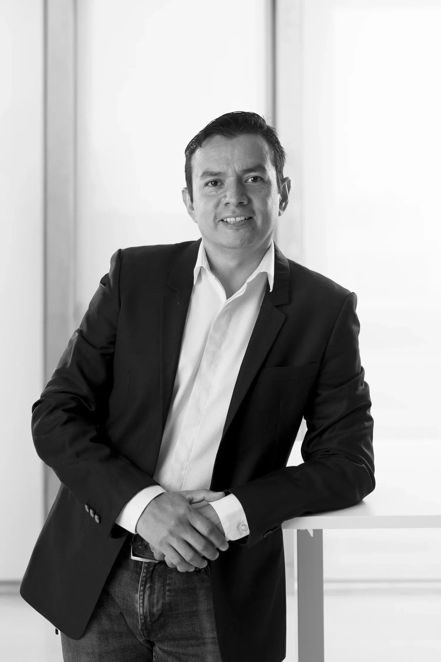 A black and white photograph of Carlos Campos in business attire, wearing a dark blazer and a white shirt, standing indoors by a white surface with a bright background.