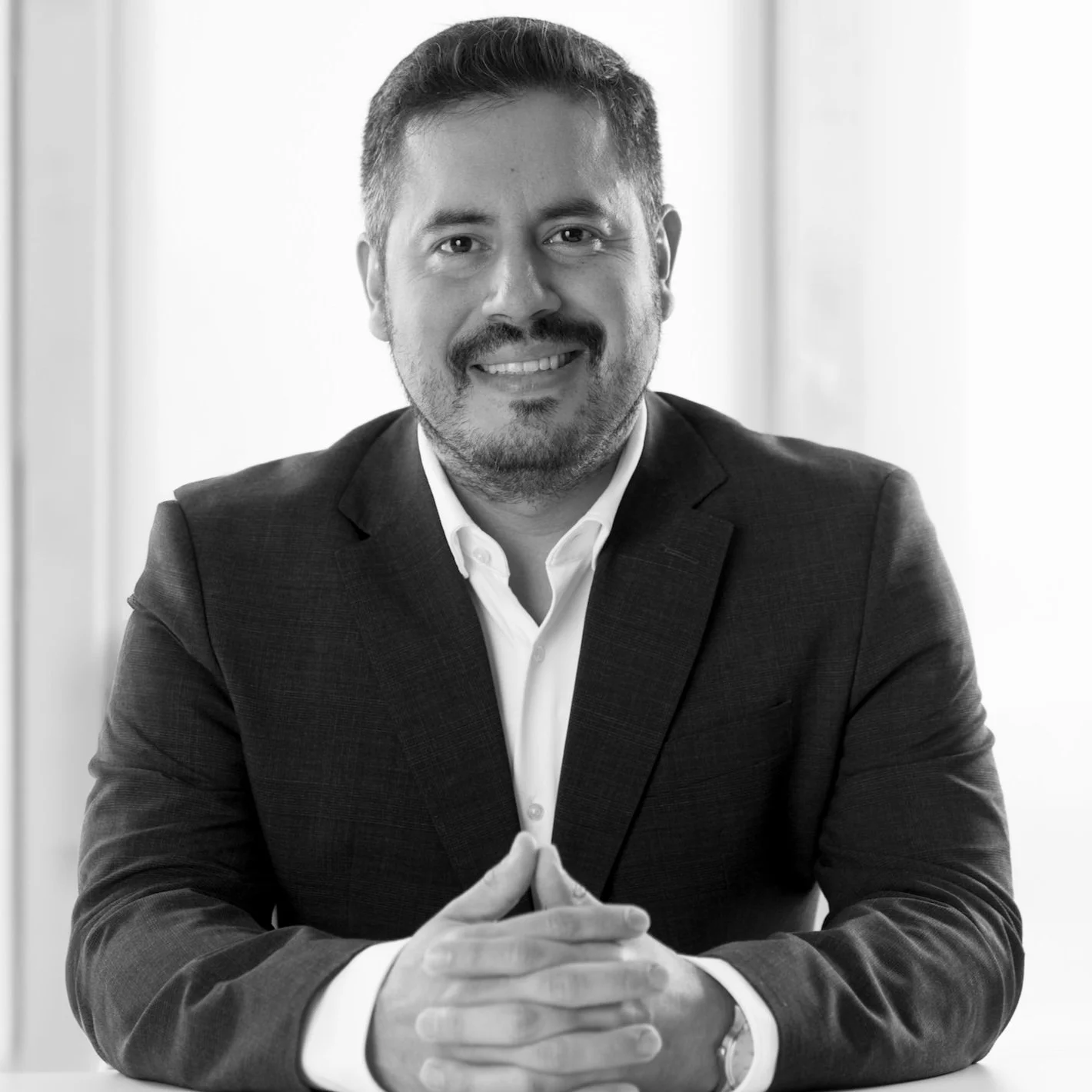 Black and white portrait of César Niebla wearing a suit, smiling, with hands clasped on a table.