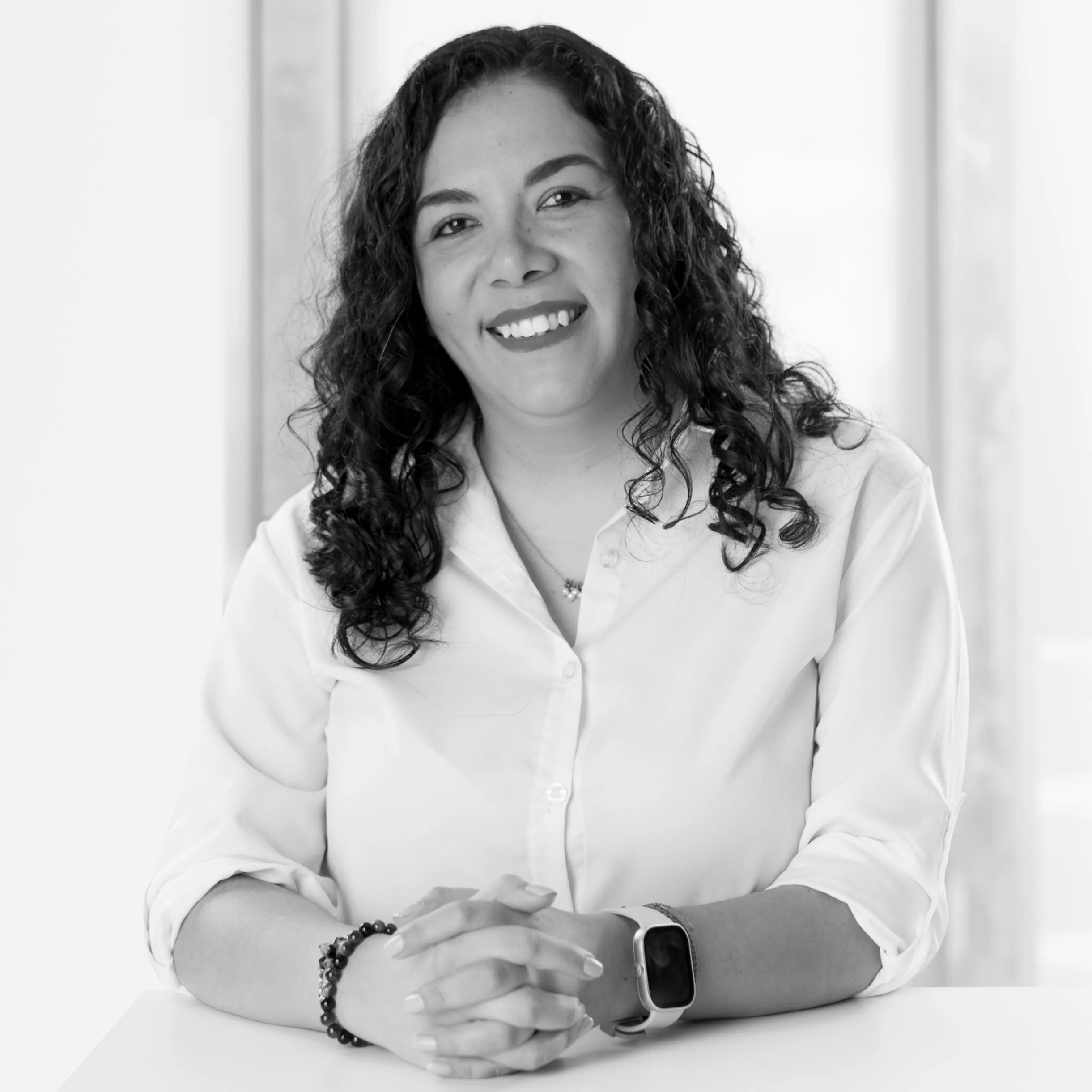 Black and white portrait of Evelyn Sanchez with curly hair, wearing a white shirt, smiling, with her hands clasped on a table, in front of a neutral background.