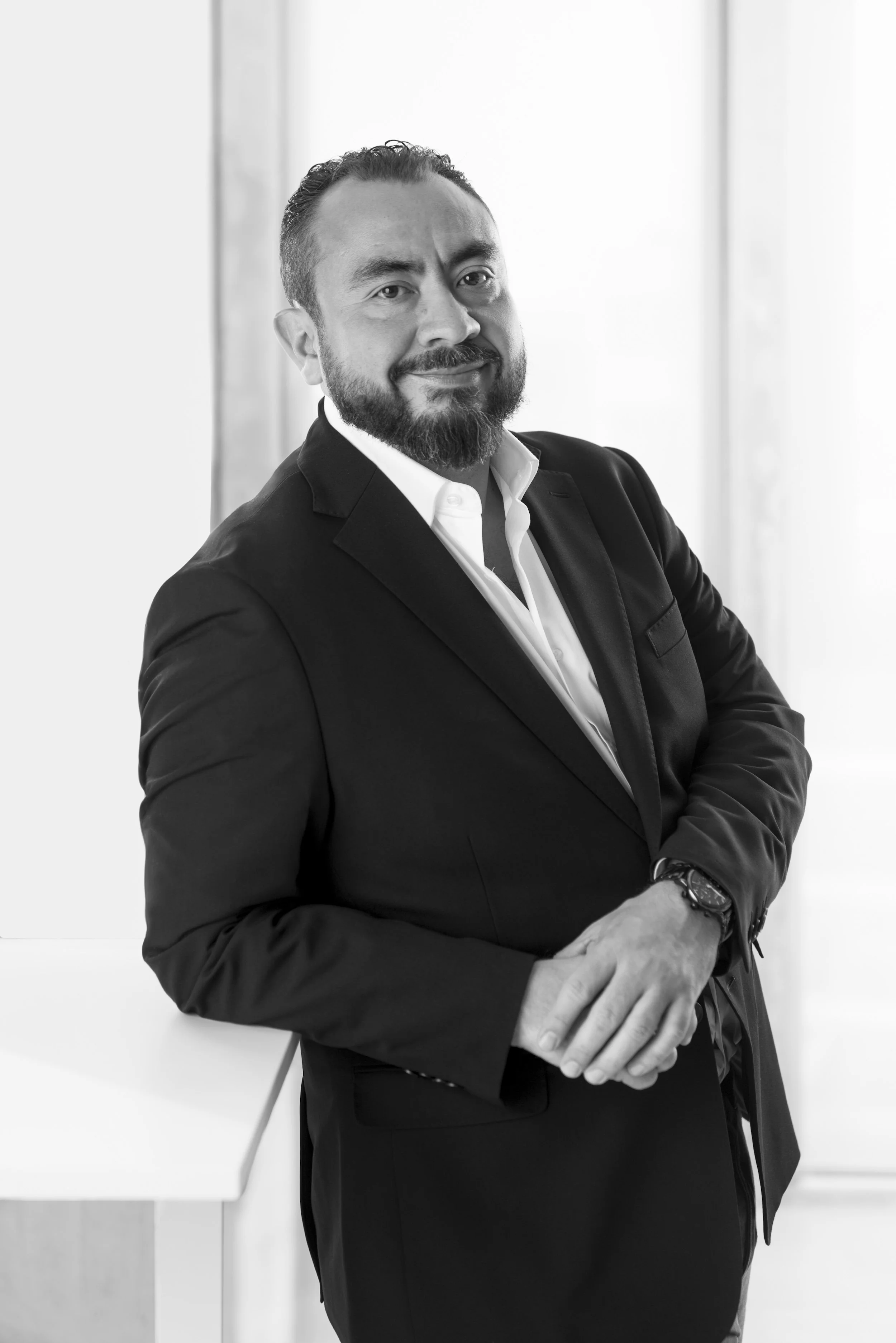 Black and white photo of Gerardo Silva in a suit standing with his arms crossed, smiling slightly, in front of a light-colored background.