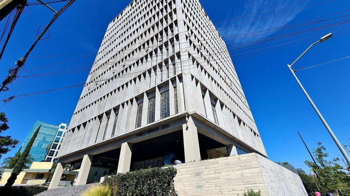 A tall modern office building with a geometric facade and large windows, located on a city street with a clear blue sky overhead.