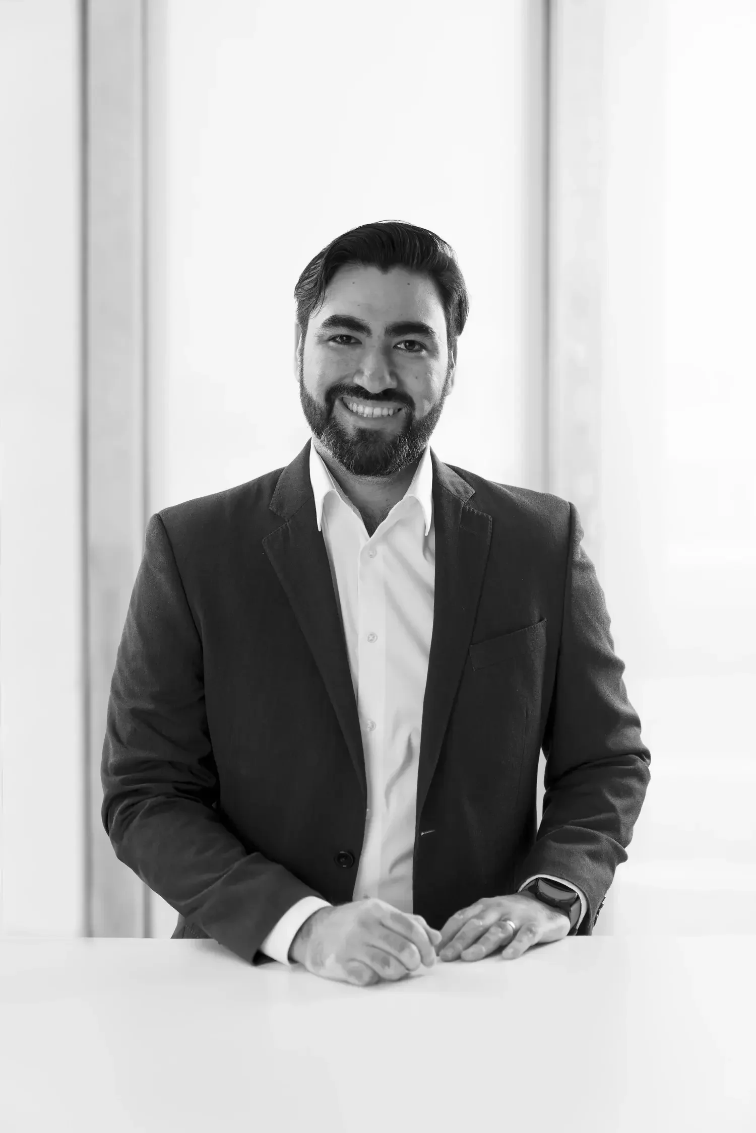 Black and white photo of Isaac Gonzalez wearing a suit and white shirt, smiling and leaning on a table.