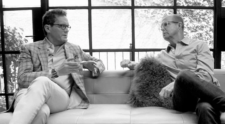 Joshua Abram and Alan Murray sitting on a couch in a bright room with large windows, having a conversation. One man is wearing glasses and a plaid blazer, holding a cup. The other man is wearing glasses and a light shirt.