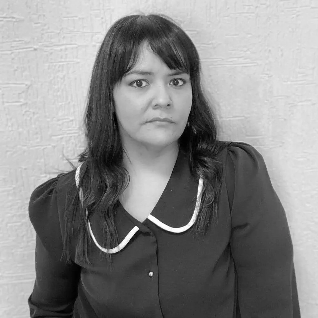 Black and white photo of Mariel Nilo, wearing a dark blouse with a white-trimmed collar, standing against a textured wall, looking directly at the camera with a serious expression.