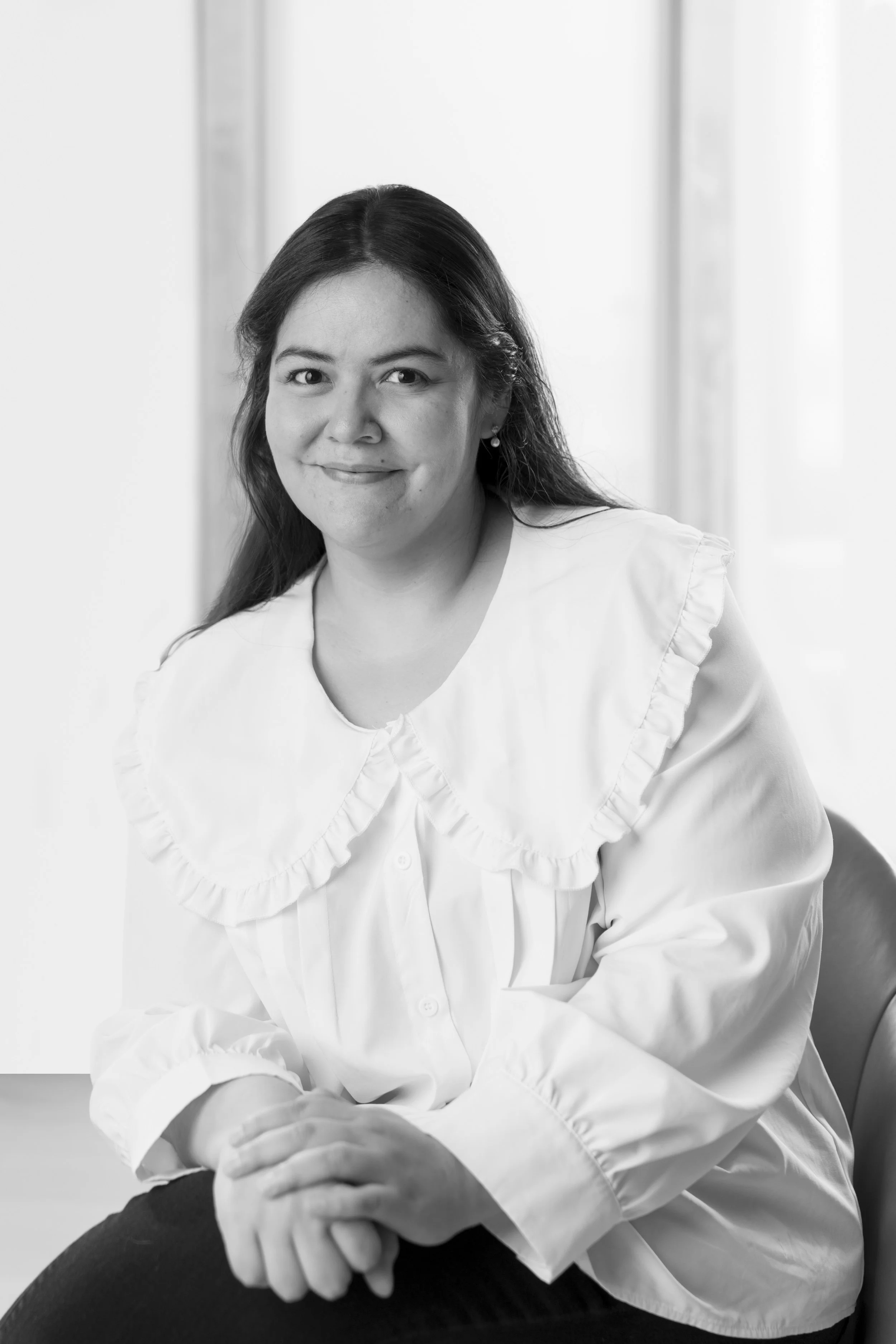 Black and white portrait of Mariel Nilo , wearing a collared blouse with ruffled edges, sitting with her hands clasped on her lap, smiling softly, against a bright background.