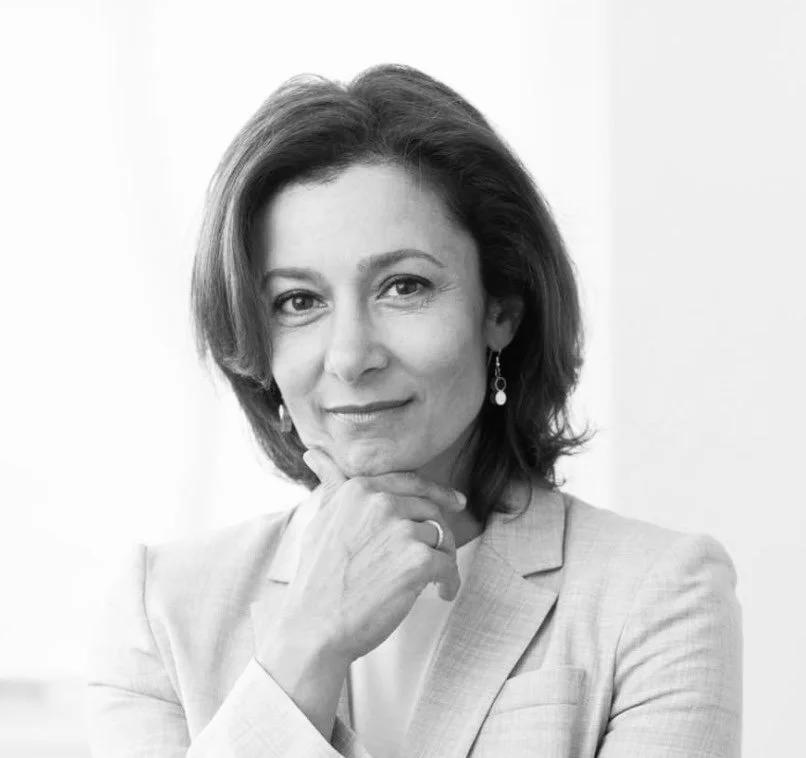 Black and white portrait of Mina Alikani with shoulder-length hair, wearing a blazer and earrings, holding her chin with her hand, looking confidently at the camera.