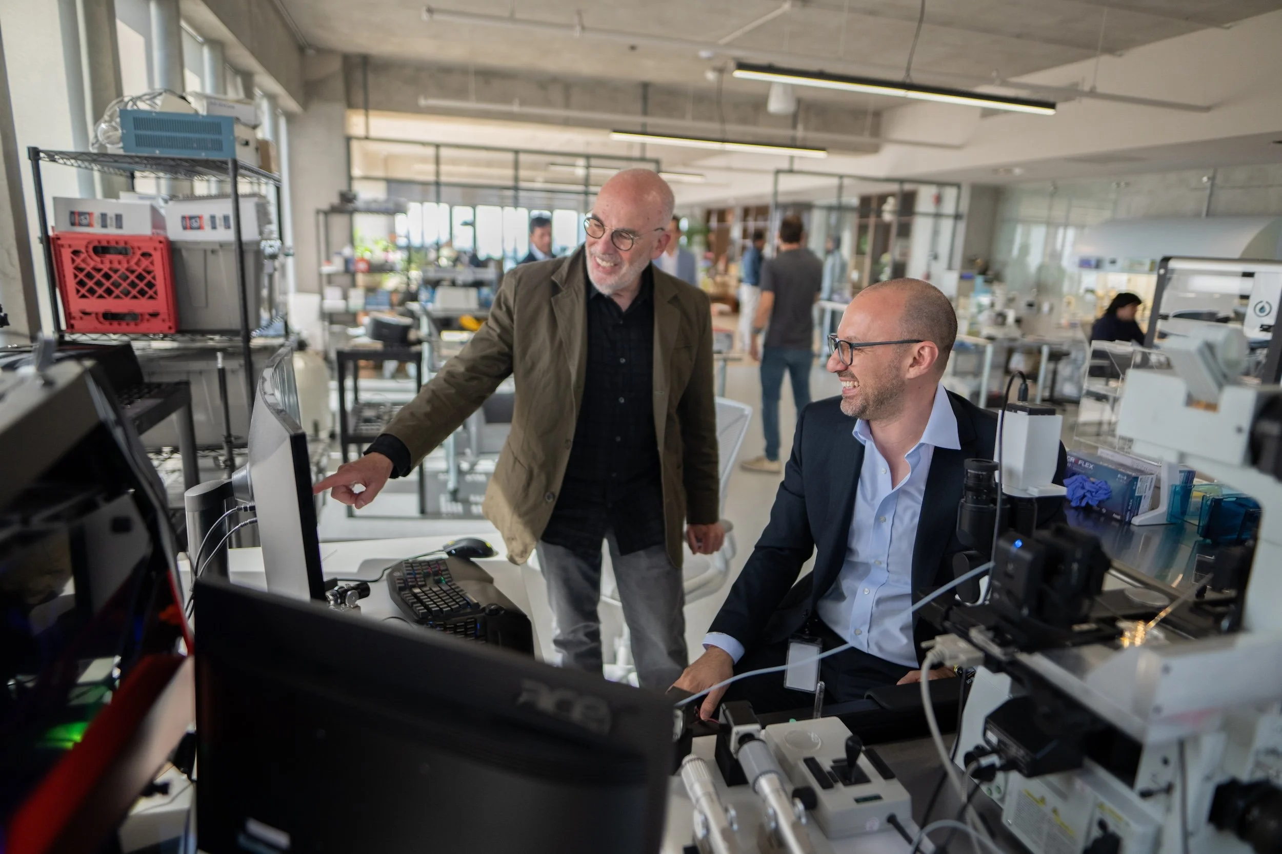 Two men in a modern office or lab, one seated at a desk with computer equipment and the other standing next to him, smiling and pointing at the screen, engaged in discussion.