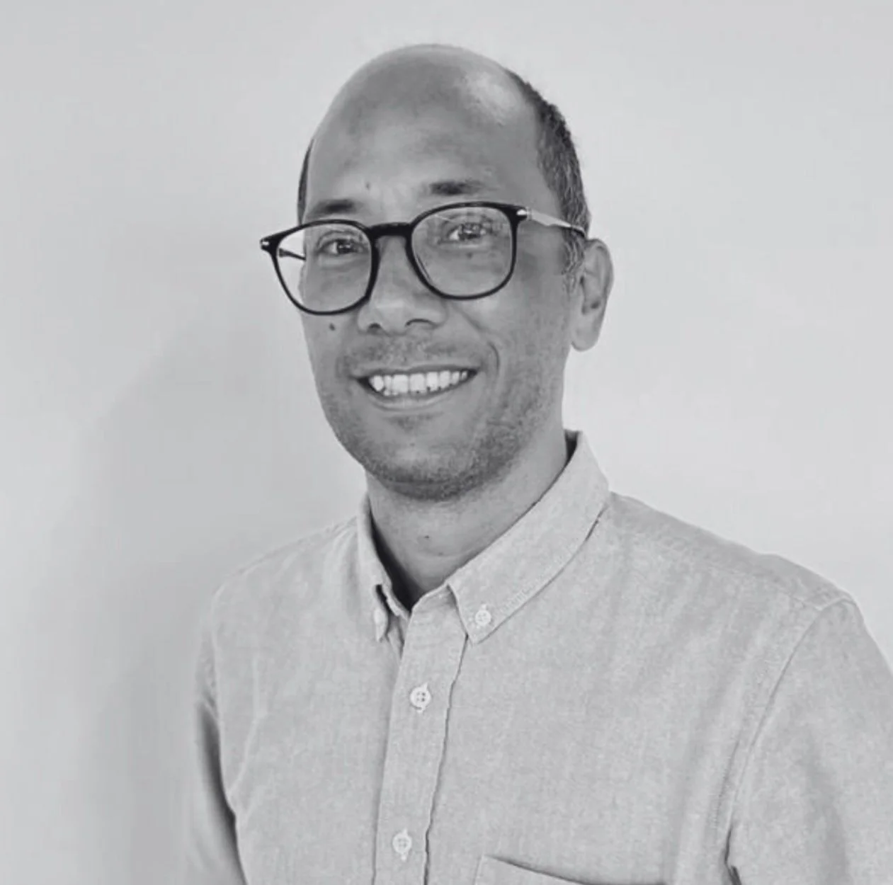 Black and white portrait of Víctor Rico wearing glasses and a button-up shirt, standing against a plain background.
