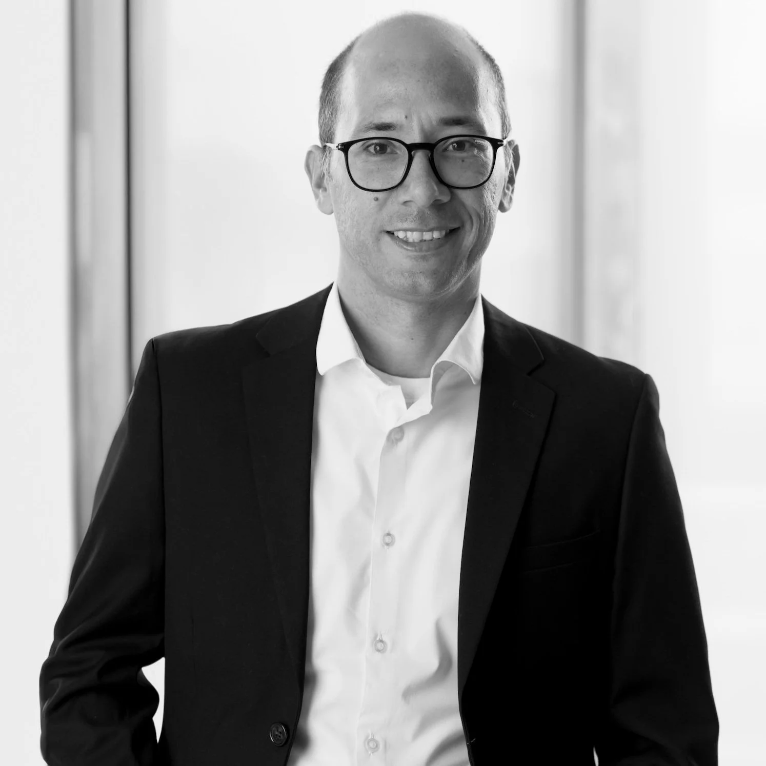 Black and white photo of Víctor Rico wearing glasses, a white shirt, and a dark suit, smiling softly in front of a light-colored background.