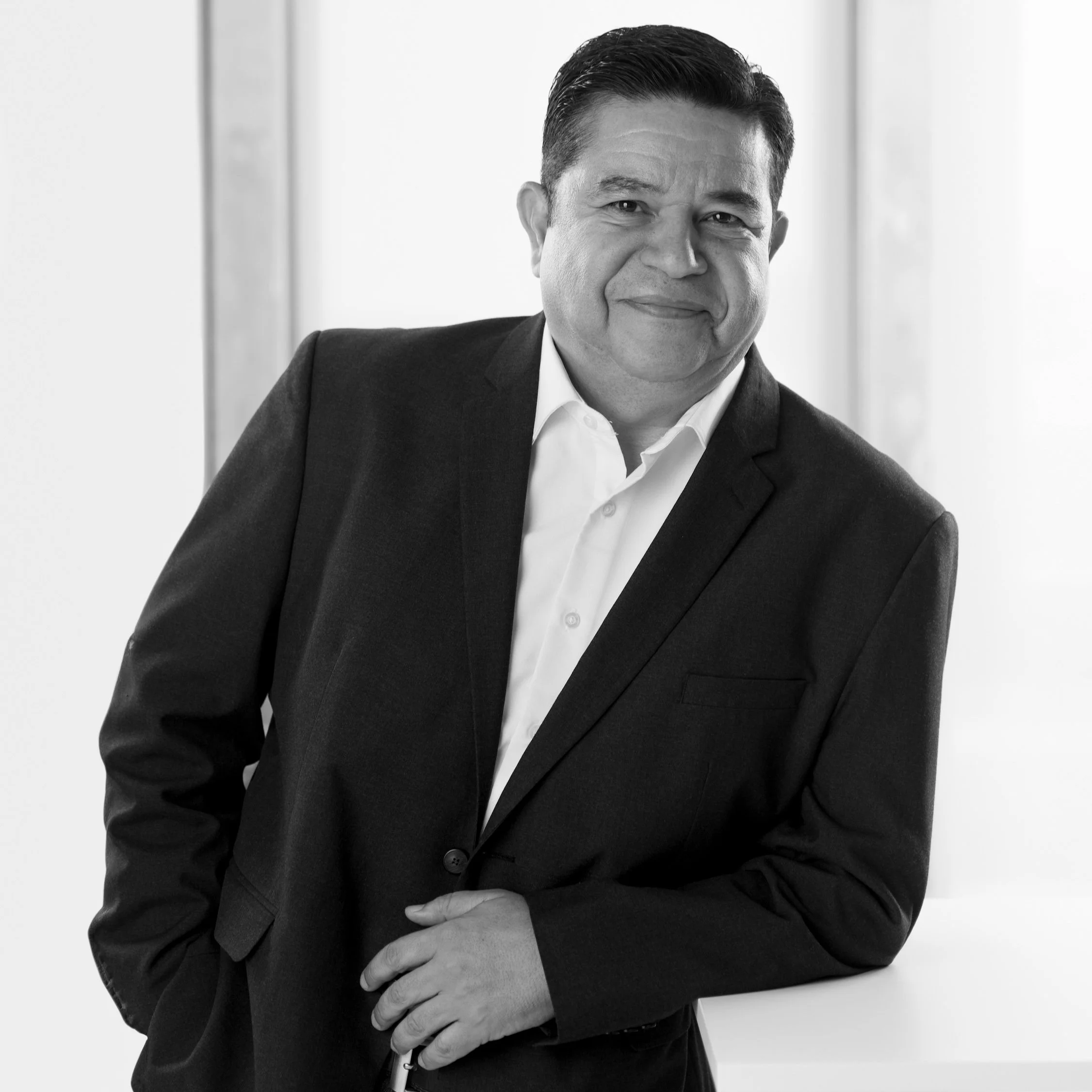 Black and white professional portrait of Vladimir Ocegueda Hernandez in a suit smiling, standing by a window.