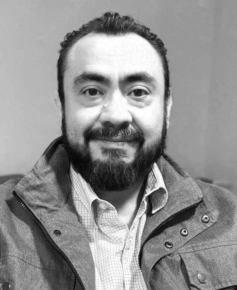 Black and white photo of Gerardo Silva in a suit standing with his arms crossed, smiling slightly, in front of a light-colored background.