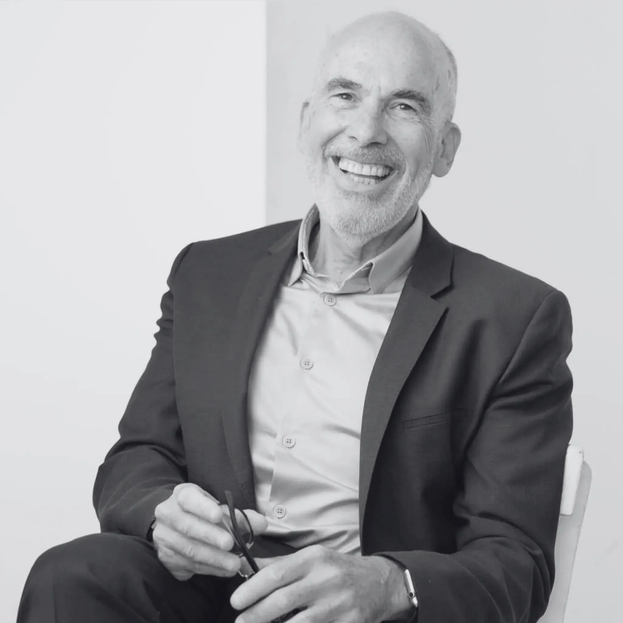 Jacques Cohen, wearing a suit jacket and a button-up shirt, sitting on a chair and holding glasses in his right hand.