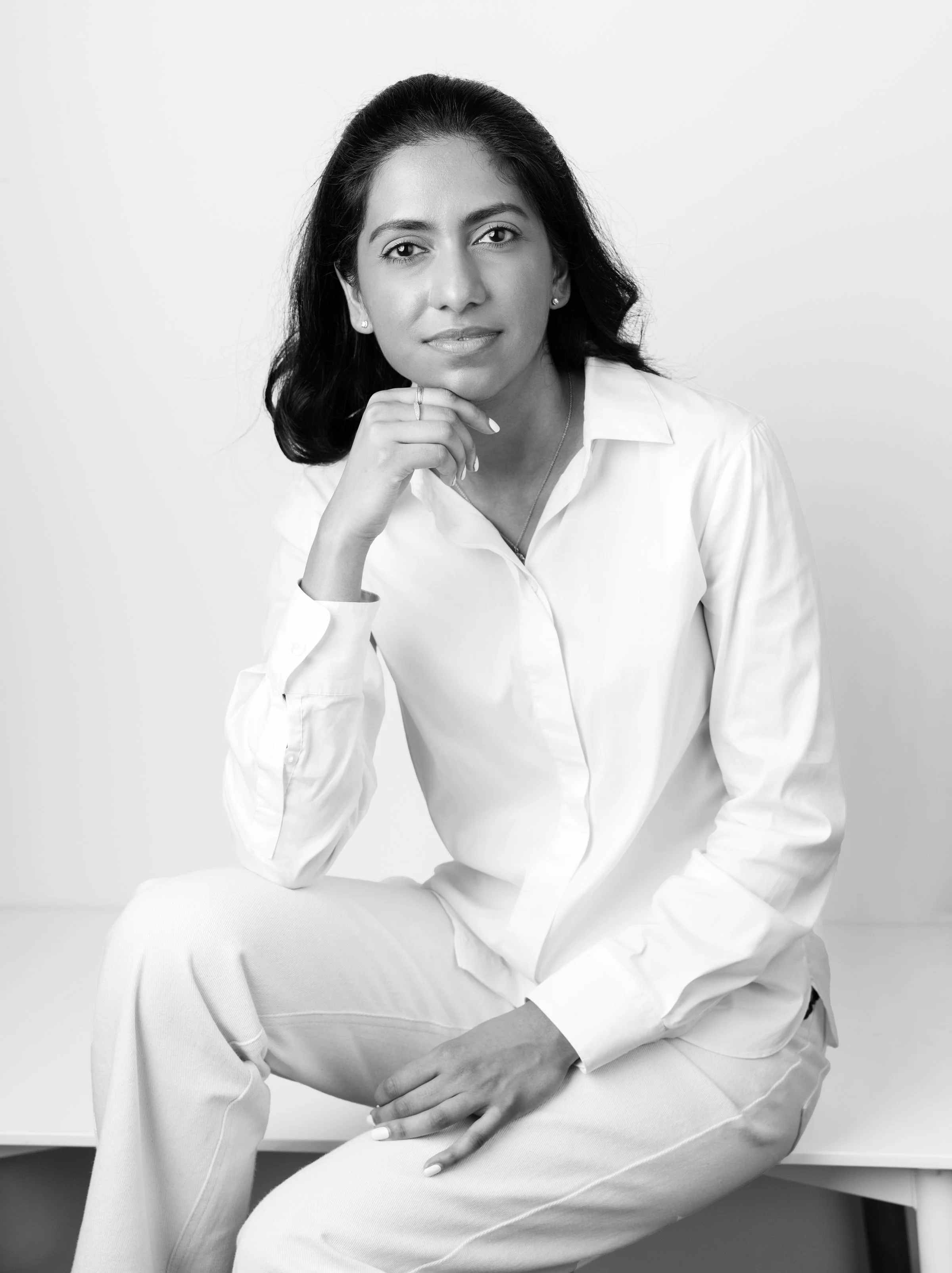 Shivani Nookala, wearing a white shirt and pants, sitting on a bench against a plain white background, looking at the camera with a neutral expression.